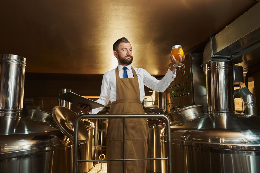 Bärtiger Brauer inspiziert in einer Brauerei ein frisch eingeschenktes Bier